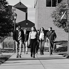Group of students walking on a sidewalk on campus with large brick buildings in the background.