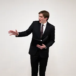 male student presenter in a suit speaks while holding a tablet, addressing an audience against a plain background