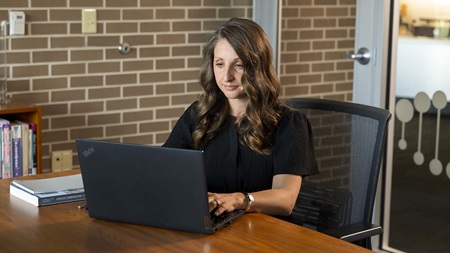 Female graduate business student sits at table working on laptop with books next to it.