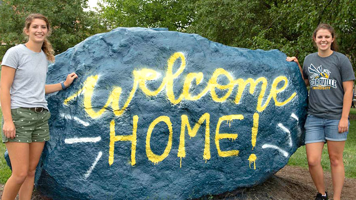 Two female students next to the rock painted with the words 'Welcome Home'.
