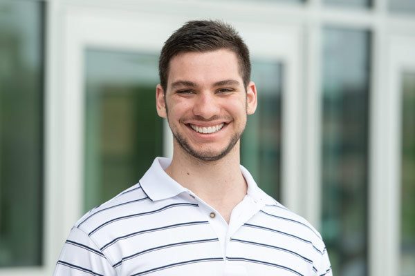 Photo of student Joseph Haire wearing a white golf shirt and smiling outside of a building.