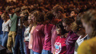 Large audience of students standing with heads bowed in prayer.