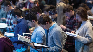 Students in Chapel standing, bibles open, in response to the reading of God's Word.