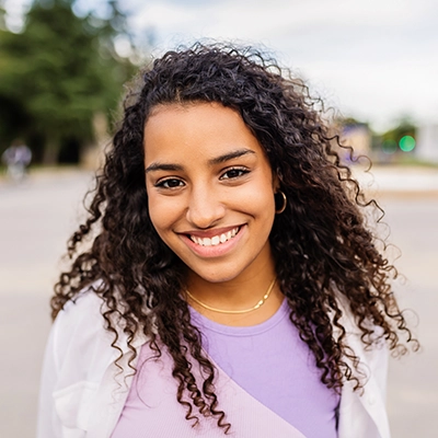 Confident student smiling outdoors, wearing a lavender top and white outer layer, with curly hair and a tree-lined background.
