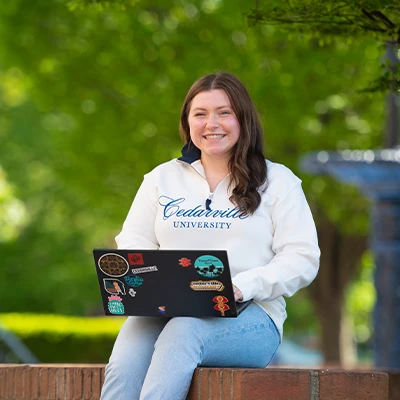 Cedarville University student outdoors with laptop, smiling on campus lawn