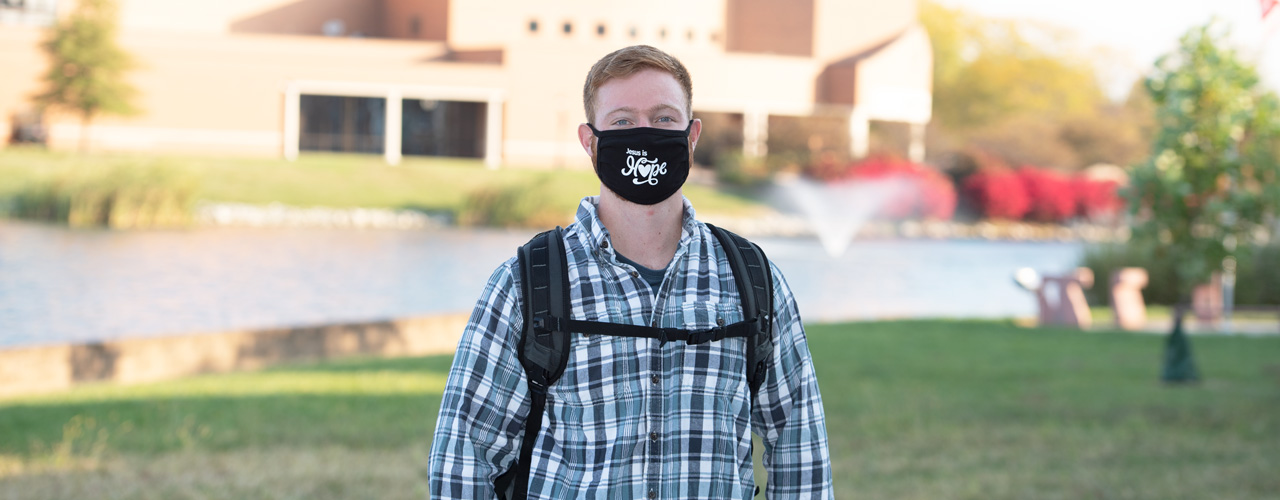 Male student in mask with Cedar Lake and Dixon Ministry Center behind him