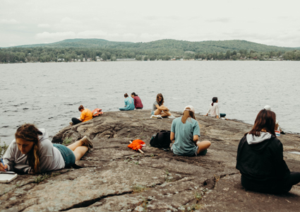 A bunch of campers reading their Bibles on a big rock next to a lake