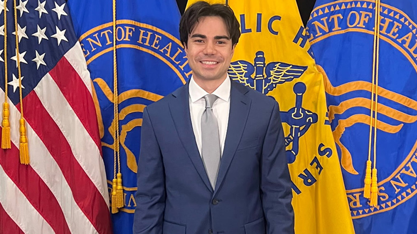 Sam Sofio standing in front of the American flag and the flag for the department of Health and Human Services flag.