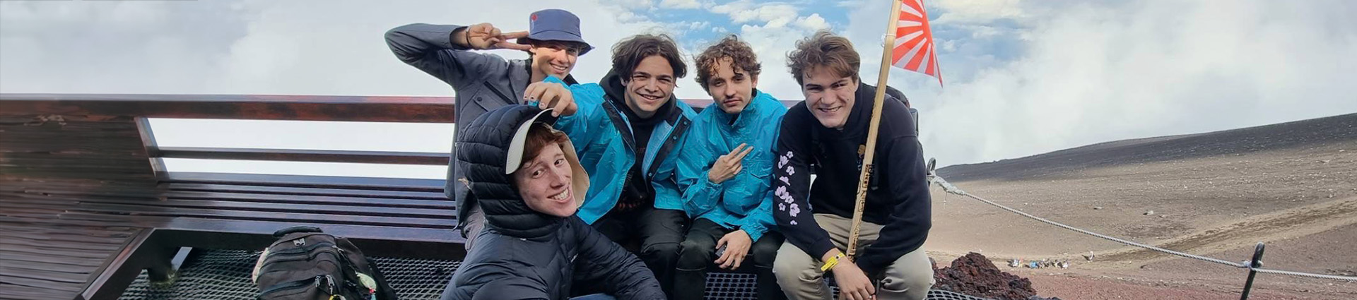 Five young men holding a small Japanese flag with sand and sky in the background.