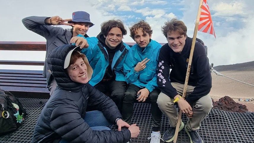 Five young men holding a small Japanese flag with sand and sky in the background.