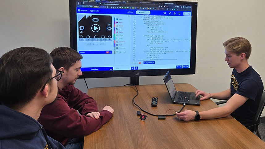 Three young men seated at table doing work on a large computer monitor.