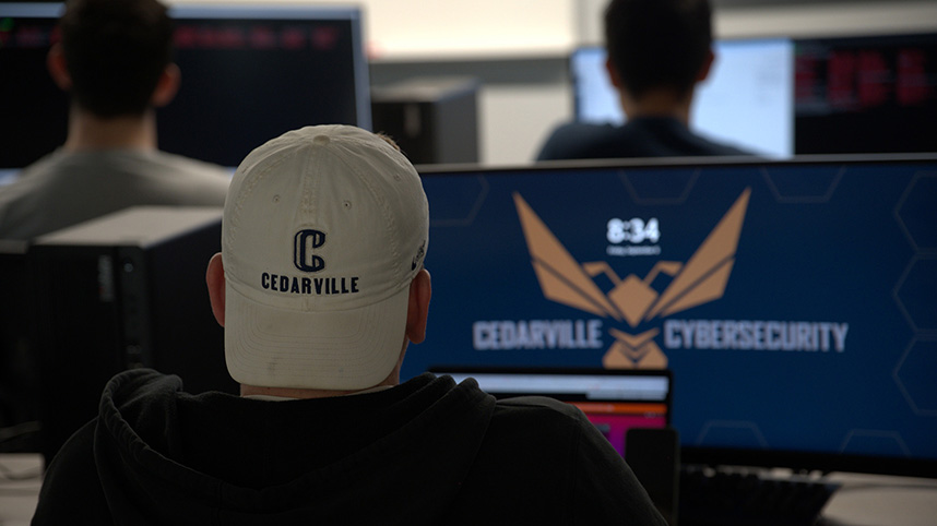 A student wearing a Cedarville University hat sits at a computer that says Cedarville Cybersecurity.