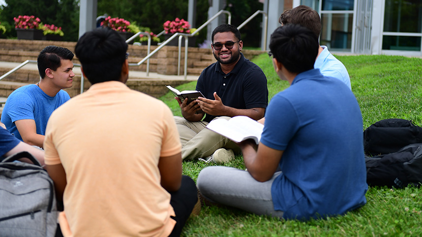 A group of male students sitting outside the Scharnberg Business and Communication Center while studying the Bible together.