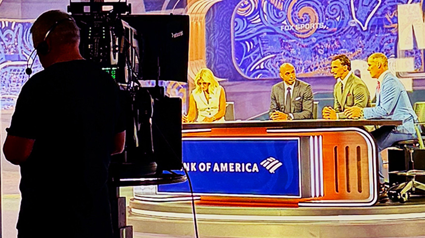 The news desk and personalities from behind a camara man in the FOX Sports studio.