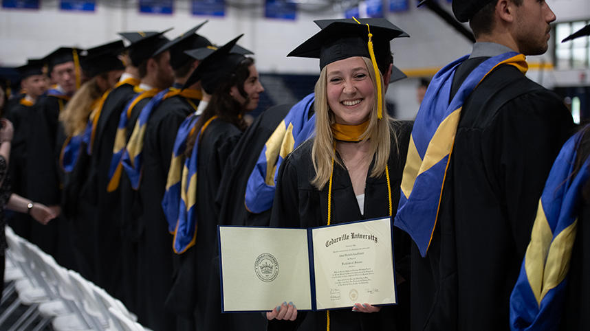 A female graduate smile while holding her diploma with other graduates in caps and gowns.