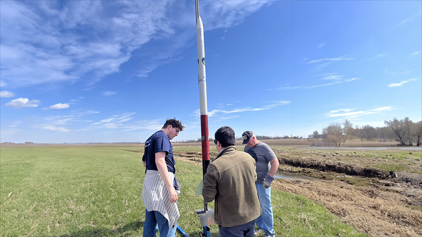 Three team members gather around the rocket before a practice launch.