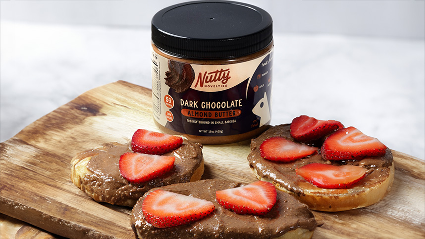 A jar of Nutty Novelties dark chocolate almond butter surrounded by toast covered in the nut butter and strawberry slices.