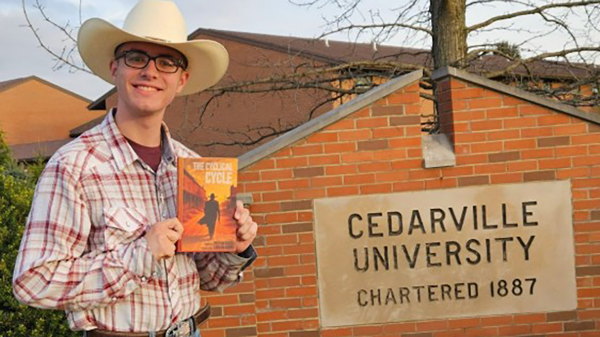 Peyton Korte in cowboy hats poses with his book by the Cedarville University sign.