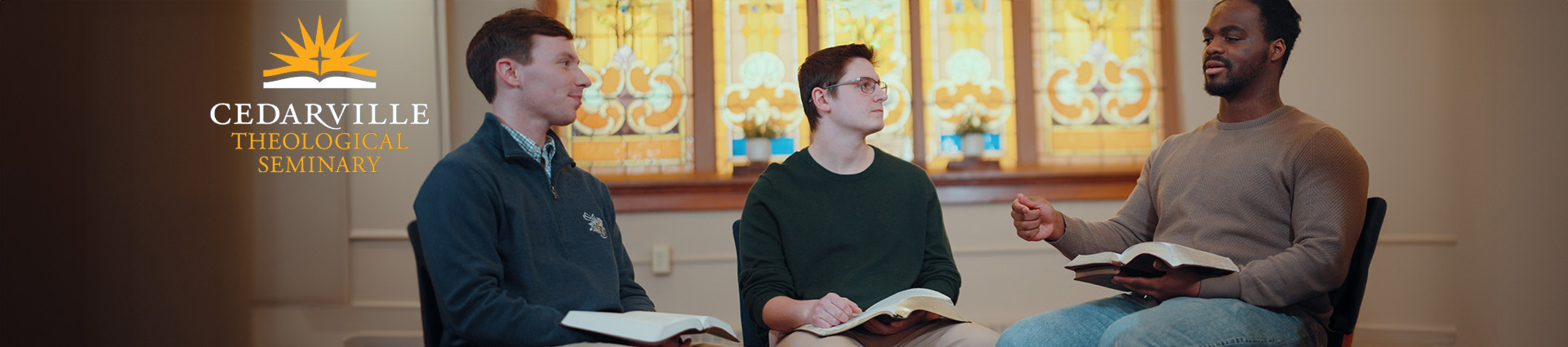 Three male students discuss the Bible while seated in front of a stained glass window.