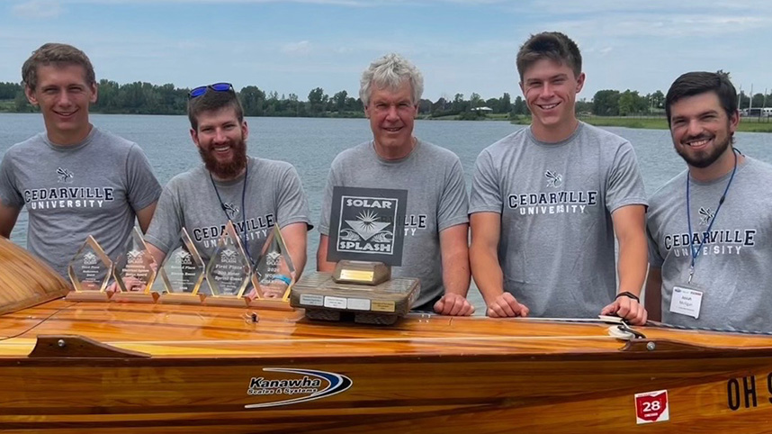 Solar Splash team of 4 students and Dr. Dewhurst with trophies sitting on the boat.