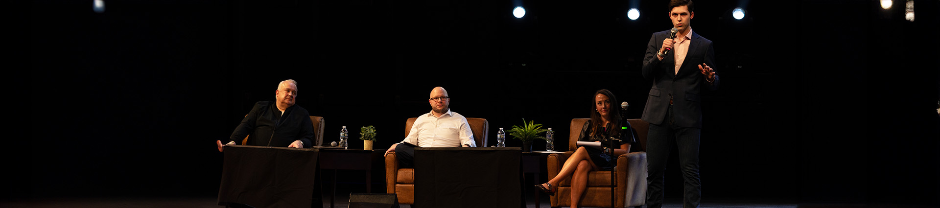 Two men and one woman sit in chairs as judges as a male student pitches his product for their endoresment.
