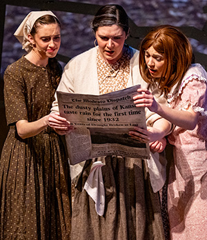 Actresses dressed in 1930s era costumes read a newspaper.