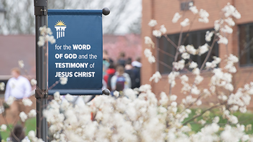 Lamp post banner that says "For the Word of God and the testimony of Jesus Christ" with spring flowers in the foreground.