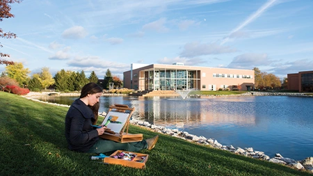 College student sitting in grass by Cedar Lake and painting the landscape