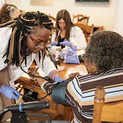 A Cedarville University pharmacy student takes an elderly woman's blood pressure in a community outreach setting, with another student assisting in the background.