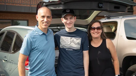 Student smiling with parents by car.