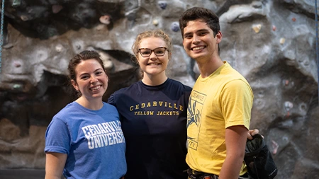 Three students smiling in front of climbing wall