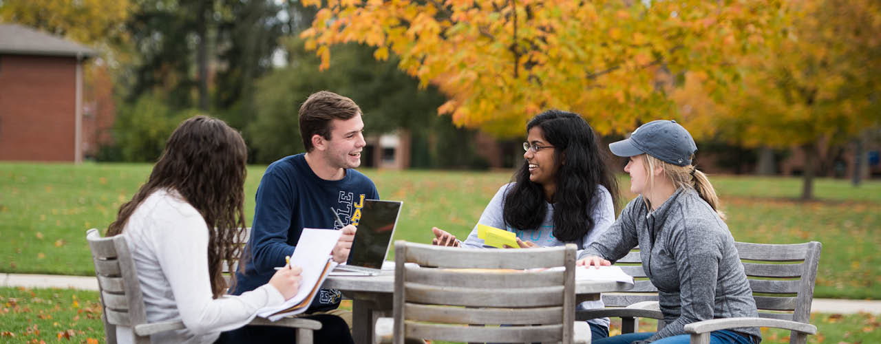 Students sitting at an outdoor table conversing with fall foliage in the background.