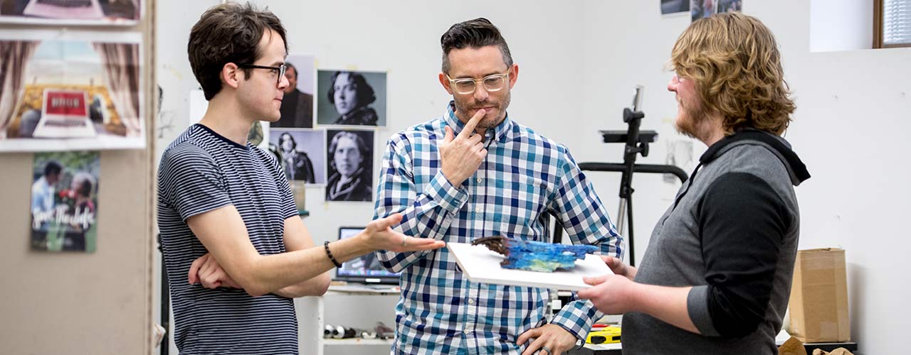 Two male art students consider a piece of art with their professor.