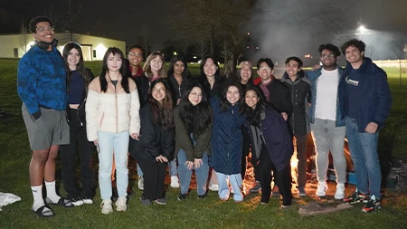 Group of sixteen students smiling in front of campfire outside
