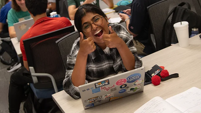 Smiling female student holding two thumbs up sitting at desk with laptop