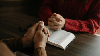 Two people praying over an open Bible on a wooden table.
