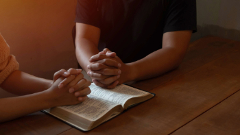 two people praying with an open Bible on a table.