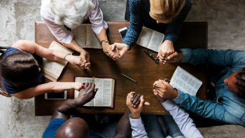 A small group holds hands in prayer around a table with their open Bibles and pens on it.