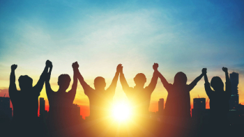 A group of people hold hands in unity overlooking a city as the sun sets.