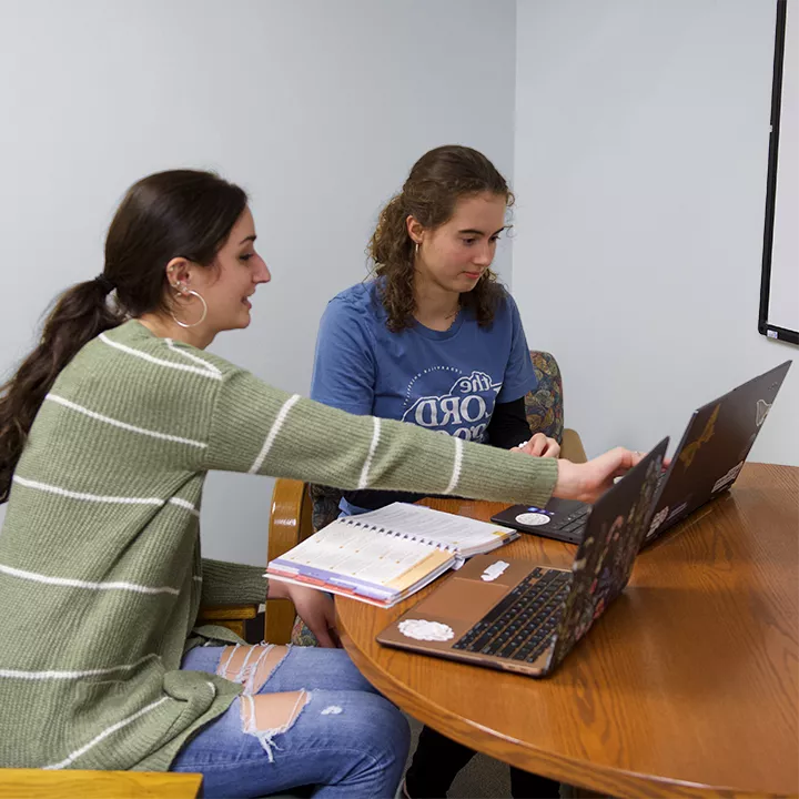 Female student tutor helping another female student using laptop computers and textbook seated at a table.