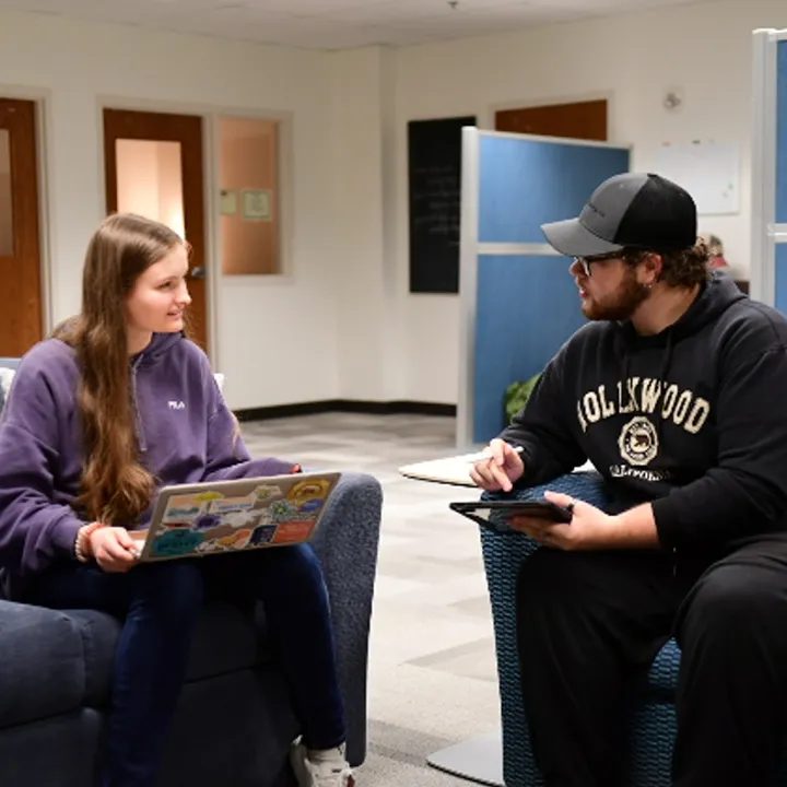 Female student tutor helping another female student using laptop computers and textbook seated at a table.
