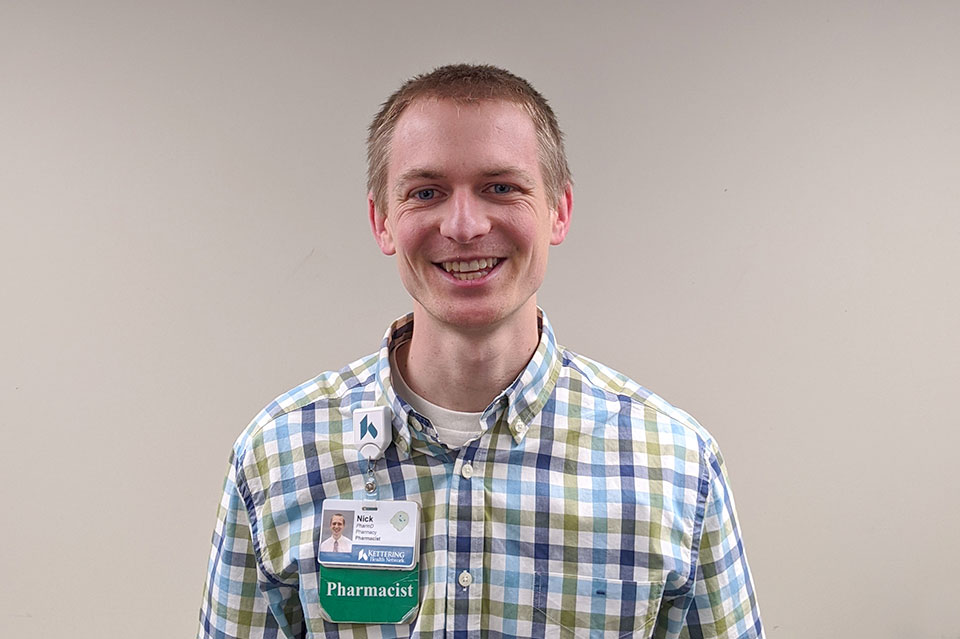 Young man standing indoors smiling, wearing a pharmacist badge.