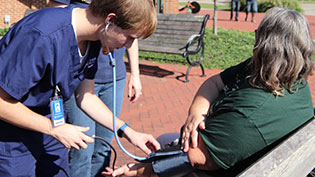 Student performing a blood pressure check for a woman sitting on a park bench.