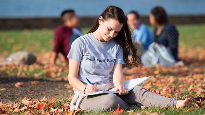 female student sitting in grass in the fall writing in a notebook