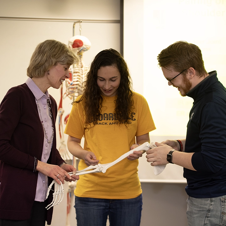 Professor and two students examine a human leg bone model in an anatomy classroom.