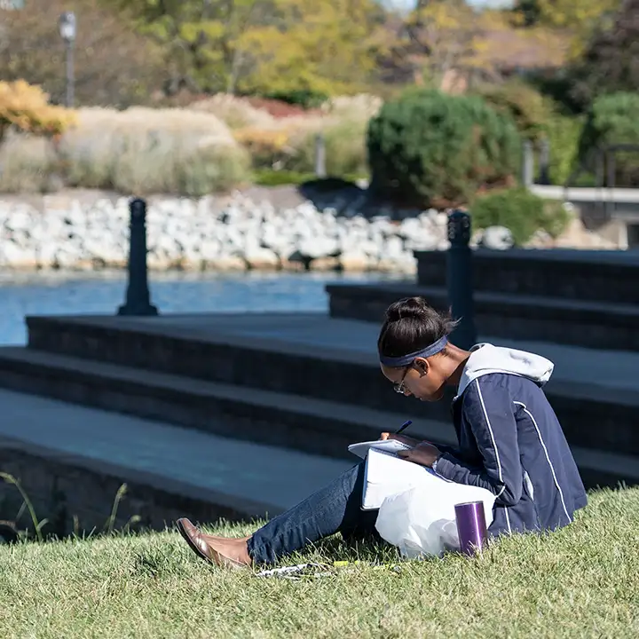 Woman sitting in the grass by the BTS steps writing in a paper notebook.