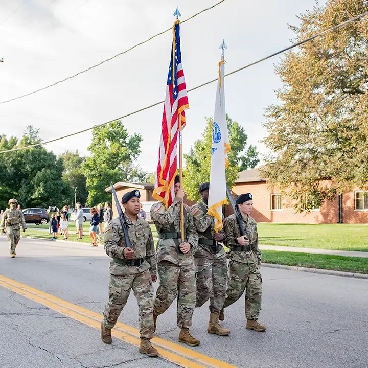 Four military personnel dressed in camouflage fatigues marching in a street parade holding flags and rifles.