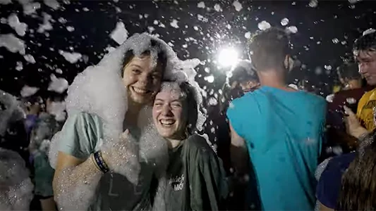 Students laughing as bubbles fall like snow all over the crowd at a nighttime event..