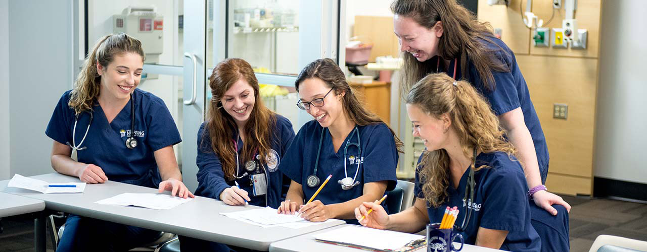Five smiling female nursing students standing and sitting behind a classroom table.