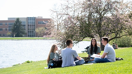 Four students reading while sitting on grass by lake